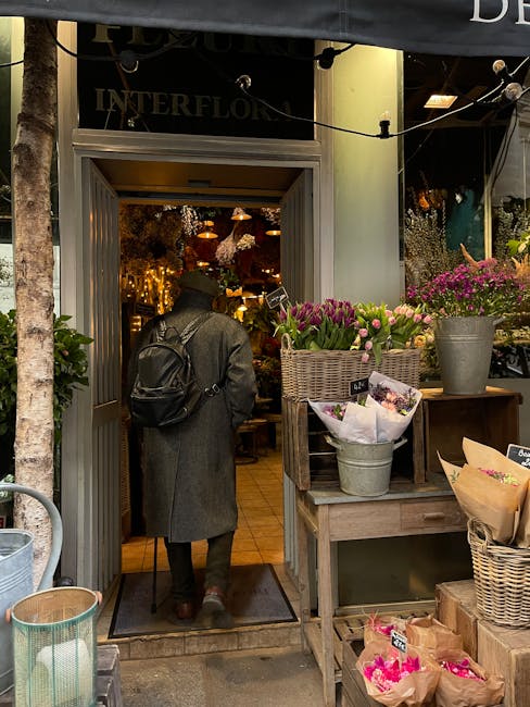 A person wearing a long grey coat and carrying a black backpack is entering a flower shop through a half-open door framed by a light grey doorframe. Outside the shop, there are various potted flowers,