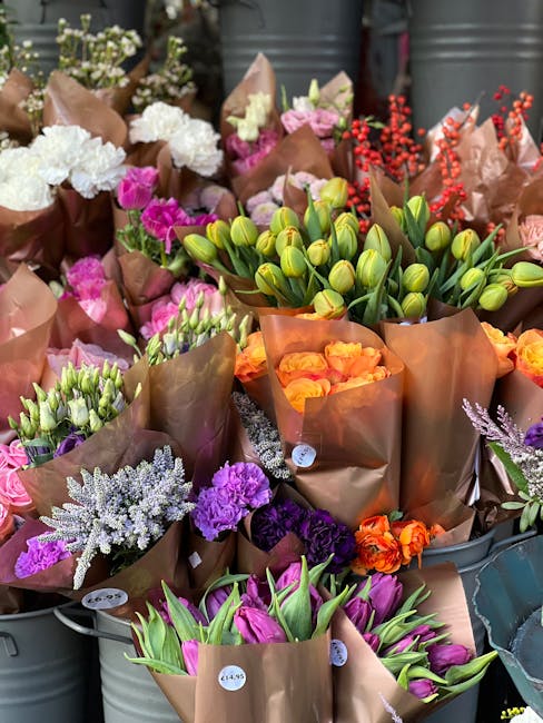 A vibrant display of fresh flower bouquets arranged in brown kraft paper wrapping, featuring a variety of blooms including white, pink, purple, and orange flowers, and green tulip buds. The bouquets a