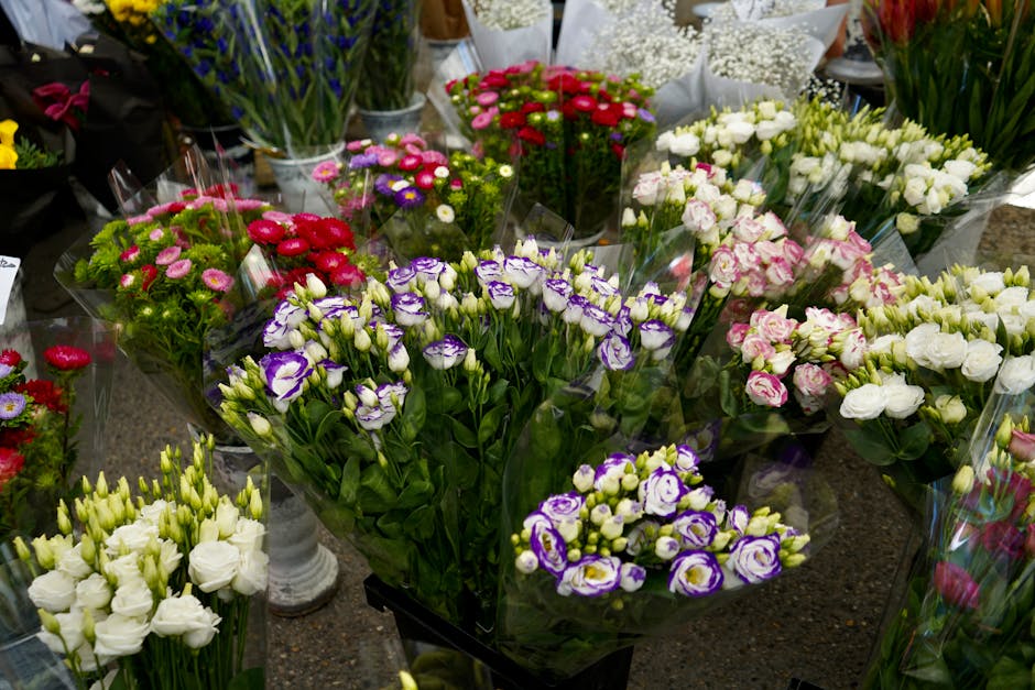 A variety of fresh floral arrangements displayed on a table, featuring tulips, roses, and lisianthus in shades of white, pink, purple, and red. The flowers are arranged in clear plastic wrapping and a
