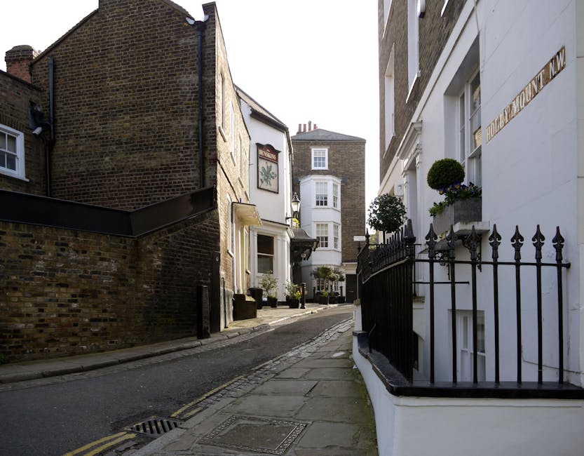 A narrow, gently sloping street in Hampstead with a mix of historic brick and white-painted buildings, featuring flower displays in window boxes and potted plants on balconies. The white building on t
