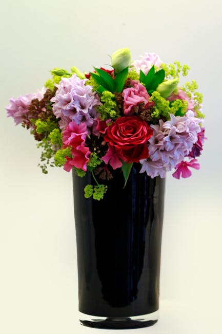 A tall, elegant black vase filled with a vibrant floral arrangement featuring a dominant single red rose at the center, surrounded by soft pink and lavender hydrangeas, white tulips, and green foliage
