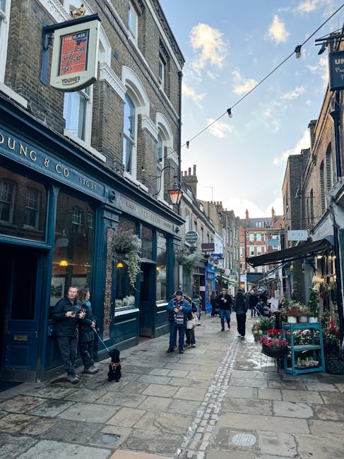 A bustling street scene featuring a floral shop with a blue facade and window display, surrounded by various storefronts and outdoor flower arrangements. Pedestrians walk along the cobblestone pavemen