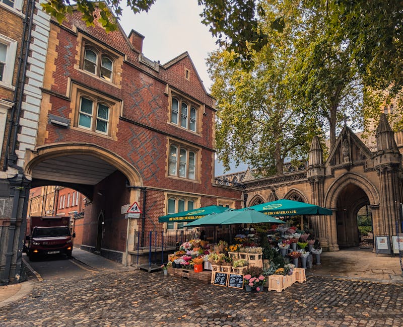 A vibrant flower stall situated on a cobbled street beneath a large green umbrella bearing the logo 'FLOWERONWELLOW.CO.UK,' displaying a colorful array of fresh flowers including pink roses, yellow tu