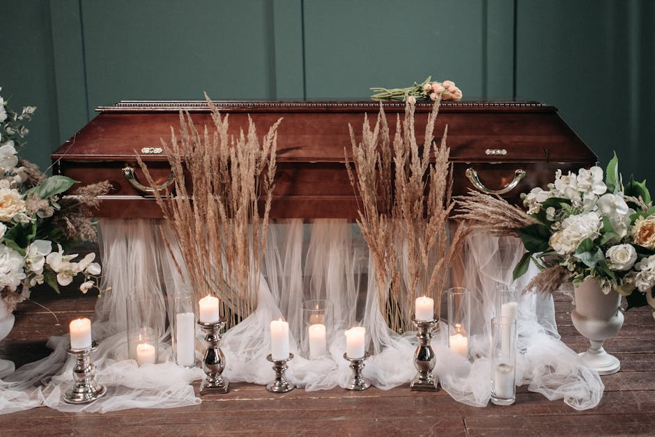 A floral arrangement displayed on a wooden floor featuring a dark wooden casket surrounded by white and pale pastel flowers in white vases, including roses and orchids. Dried pampas grass stems are pl