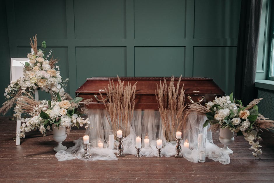 A solemn funeral floral arrangement featuring two large, elegant vases filled with a mix of white roses, hydrangeas, and other soft-colored flowers, accented with dried pampas grass and greenery. The 